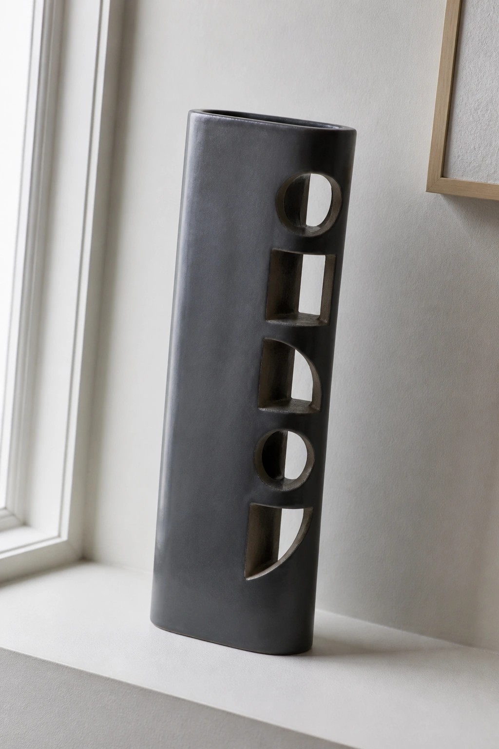 Tall dark gray ceramic vase with geometric cutouts on white windowsill
