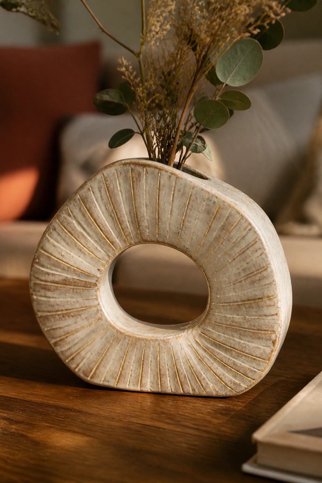 White donut-shaped ceramic vase with gold rays holding dried eucalyptus on wood