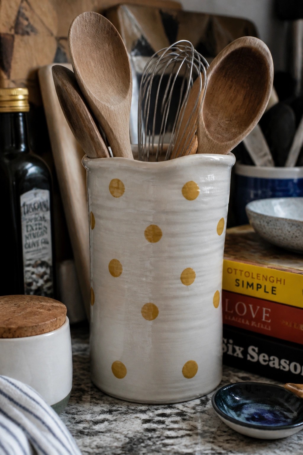 White polka-dotted ceramic crock holding wooden spoons and metal whisk on countertop