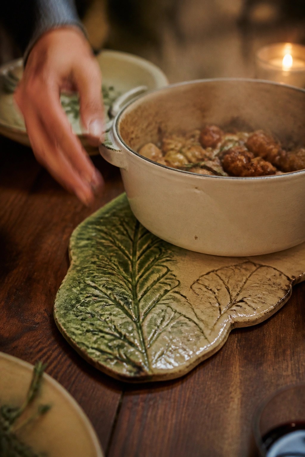 Hand reaching toward steaming pot on green leaf trivet beside candle.