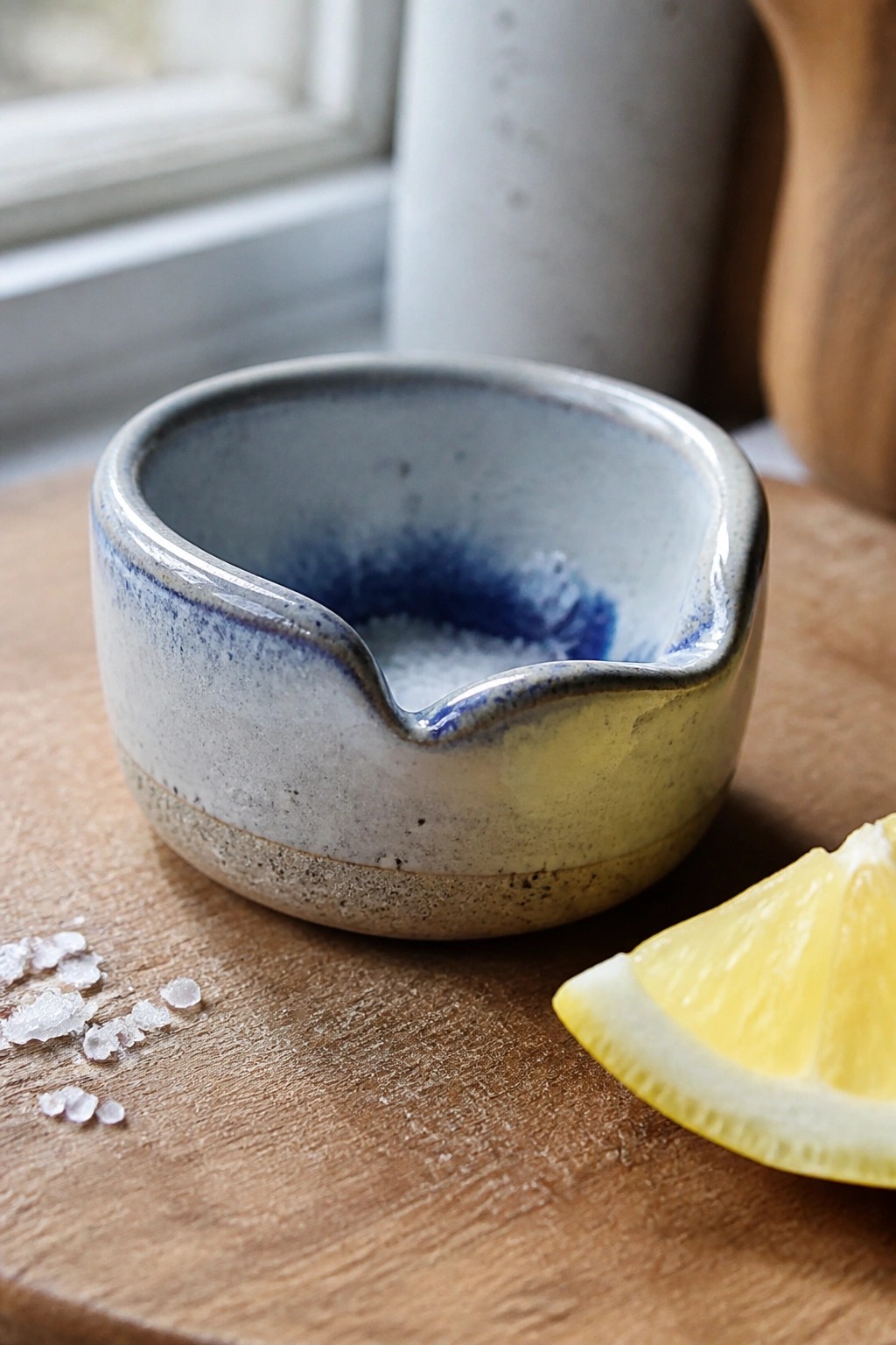 A small ceramic bowl with a spout and blue-yellow glaze sits on wood next to a lemon wedge and scattered salt.