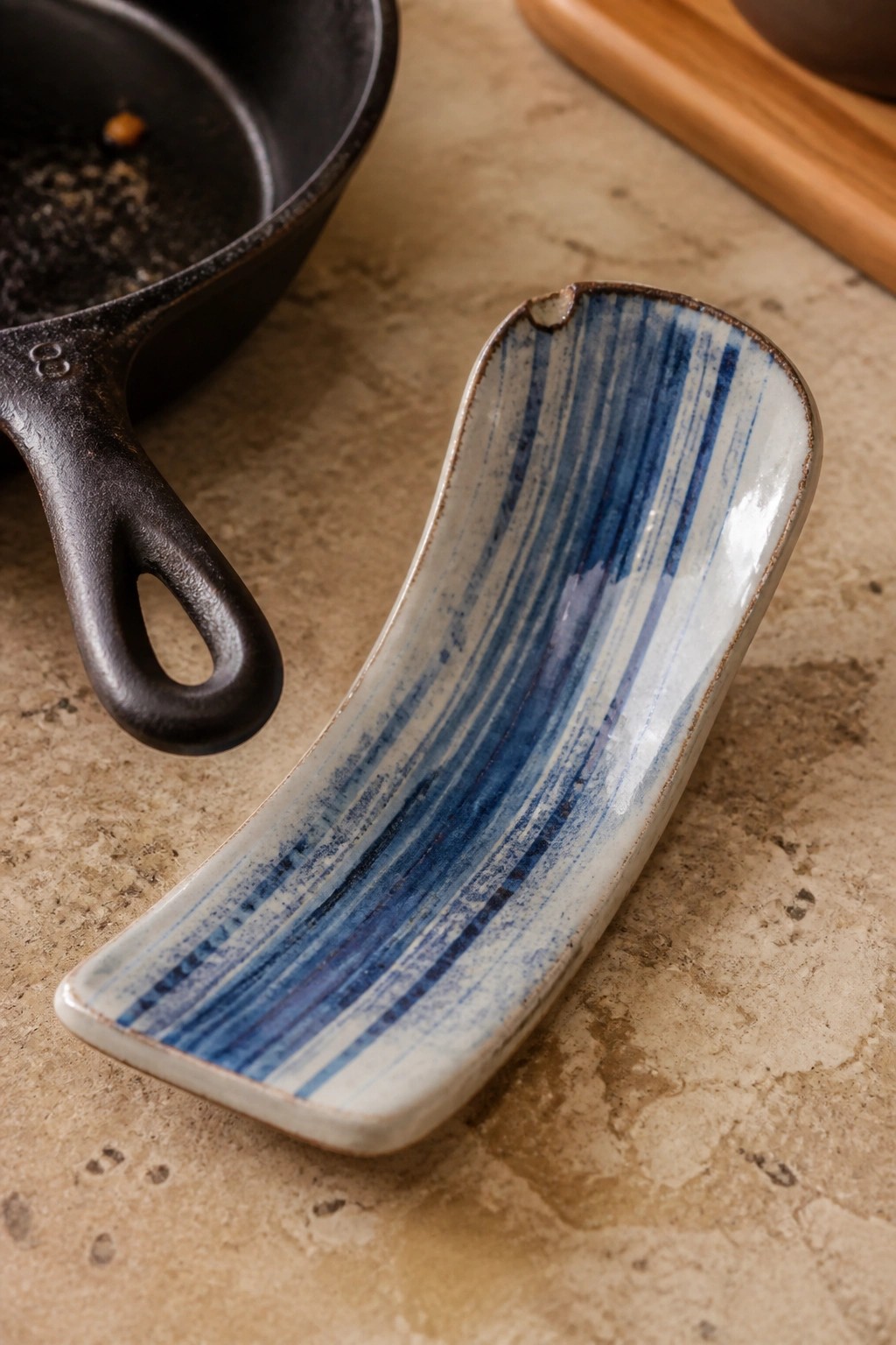 Blue striped ceramic spoon rest beside black cast iron skillet on stone counter