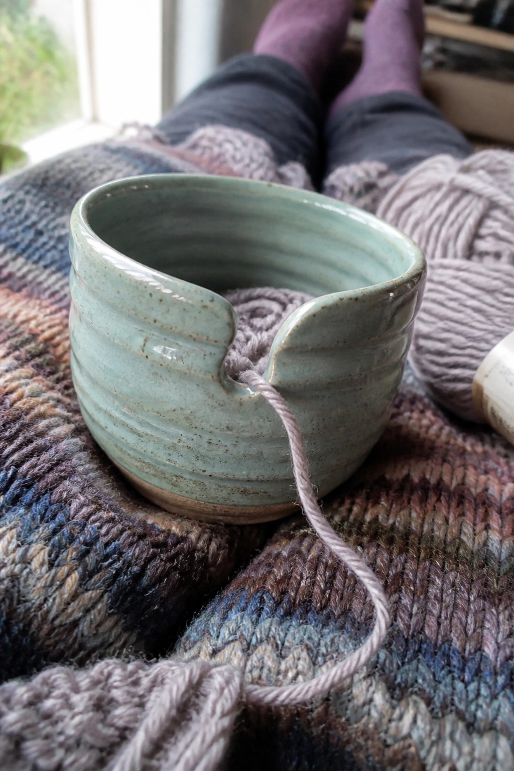 Mint green ceramic yarn bowl with purple yarn on plaid blanket near purple socks