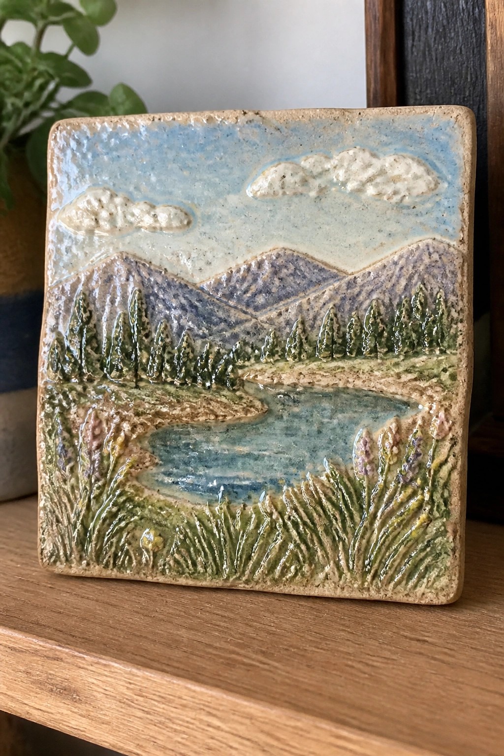 Square ceramic tile with relief landscape of mountains, pine trees, lake, and flowers.