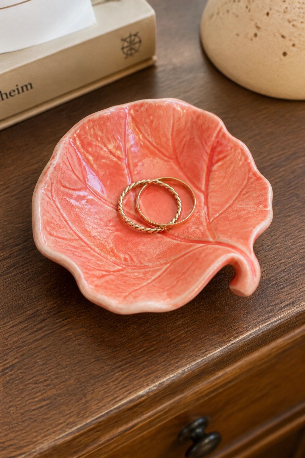 Pink leaf-shaped ceramic dish holding two gold rings on wooden table.