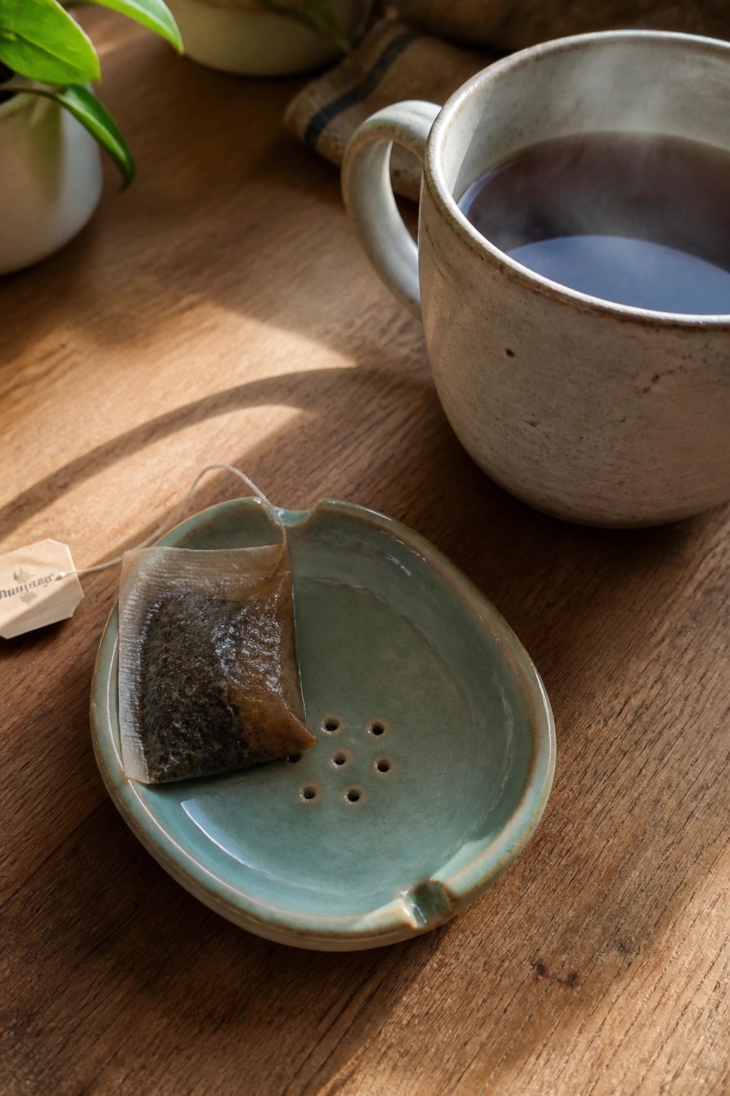 Small teal ceramic dish with drainage holes holding a tea bag beside a steaming mug on a wooden table.