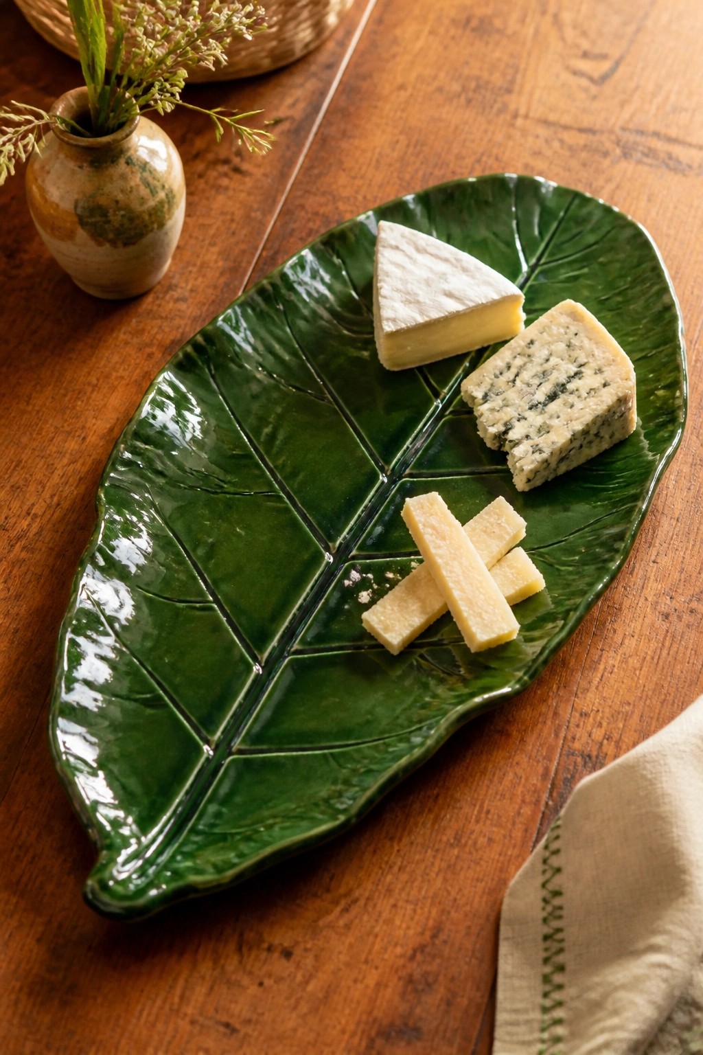 Green leaf-shaped plate with assorted cheeses on rustic wooden table beside vase