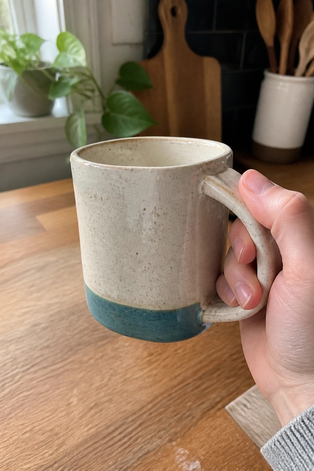 A hand holds a ceramic mug with a speckled off-white upper half and teal lower half above a wooden table.
