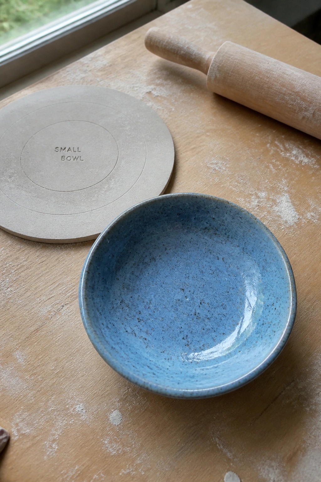 Blue speckled bowl on floured wood beside clay mold and rolling pin
