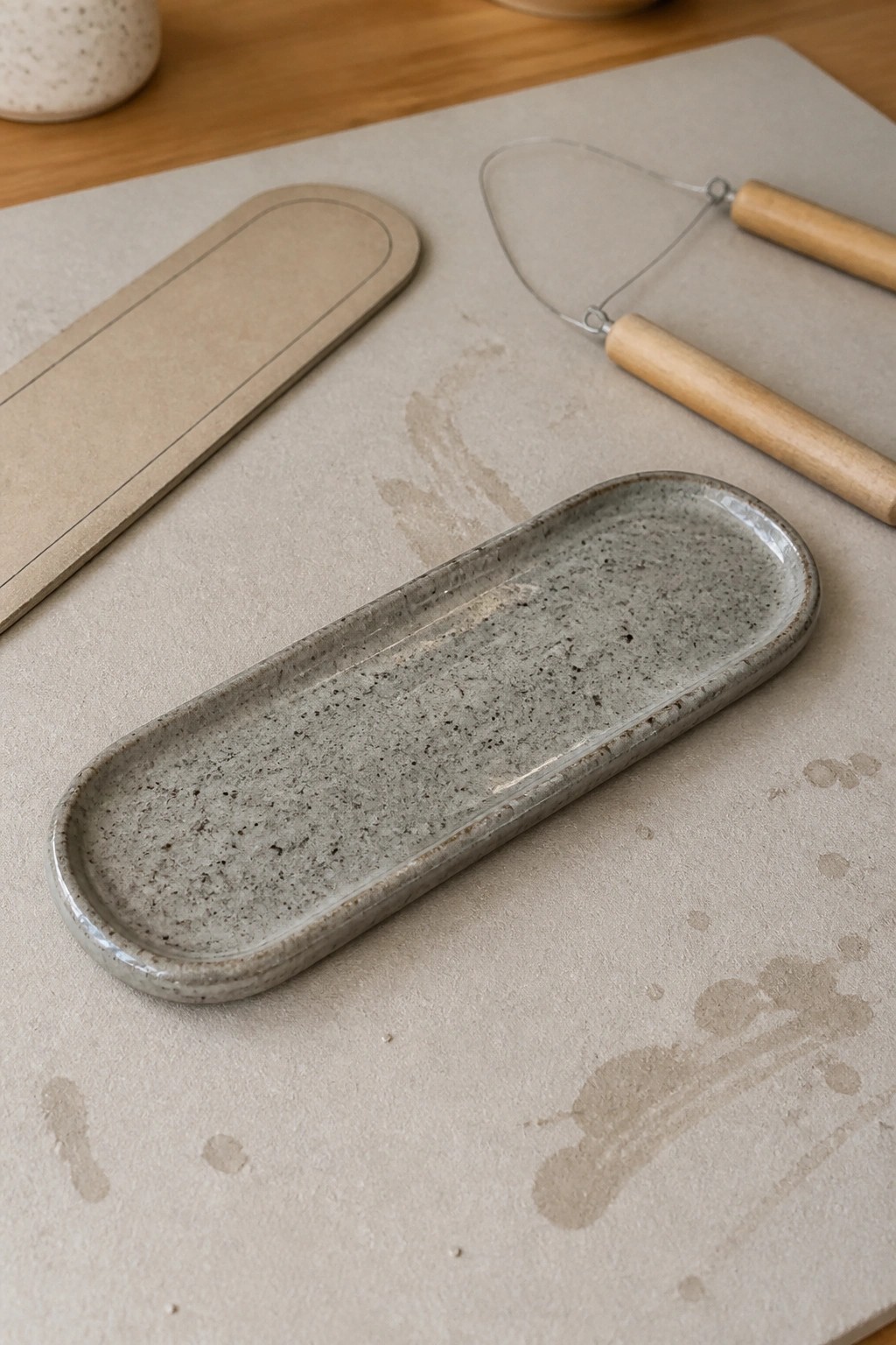 Gray speckled ceramic tray beside beige slab and wire-handled pottery tool on work surface.