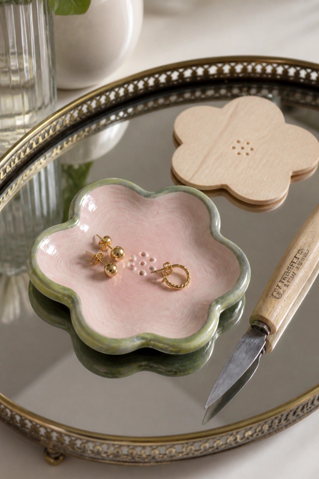 Gold earrings in pink flower-shaped ceramic dish beside wooden form and tool on tray.