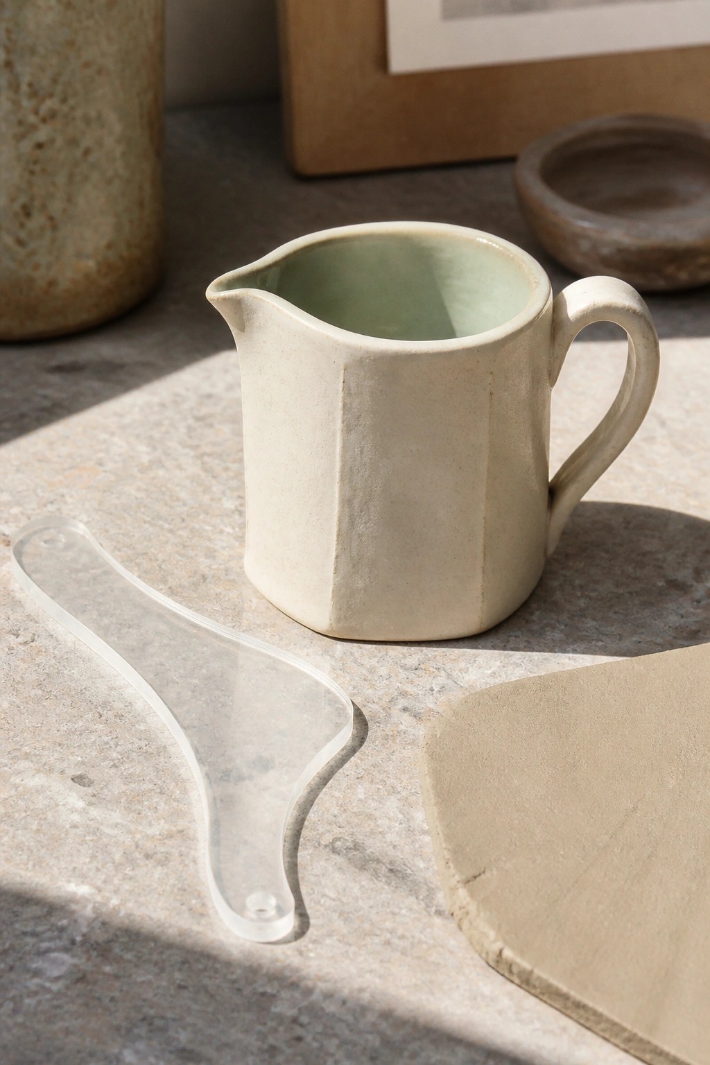 Cream ceramic pitcher with green interior beside clear pottery tool and clay on stone table