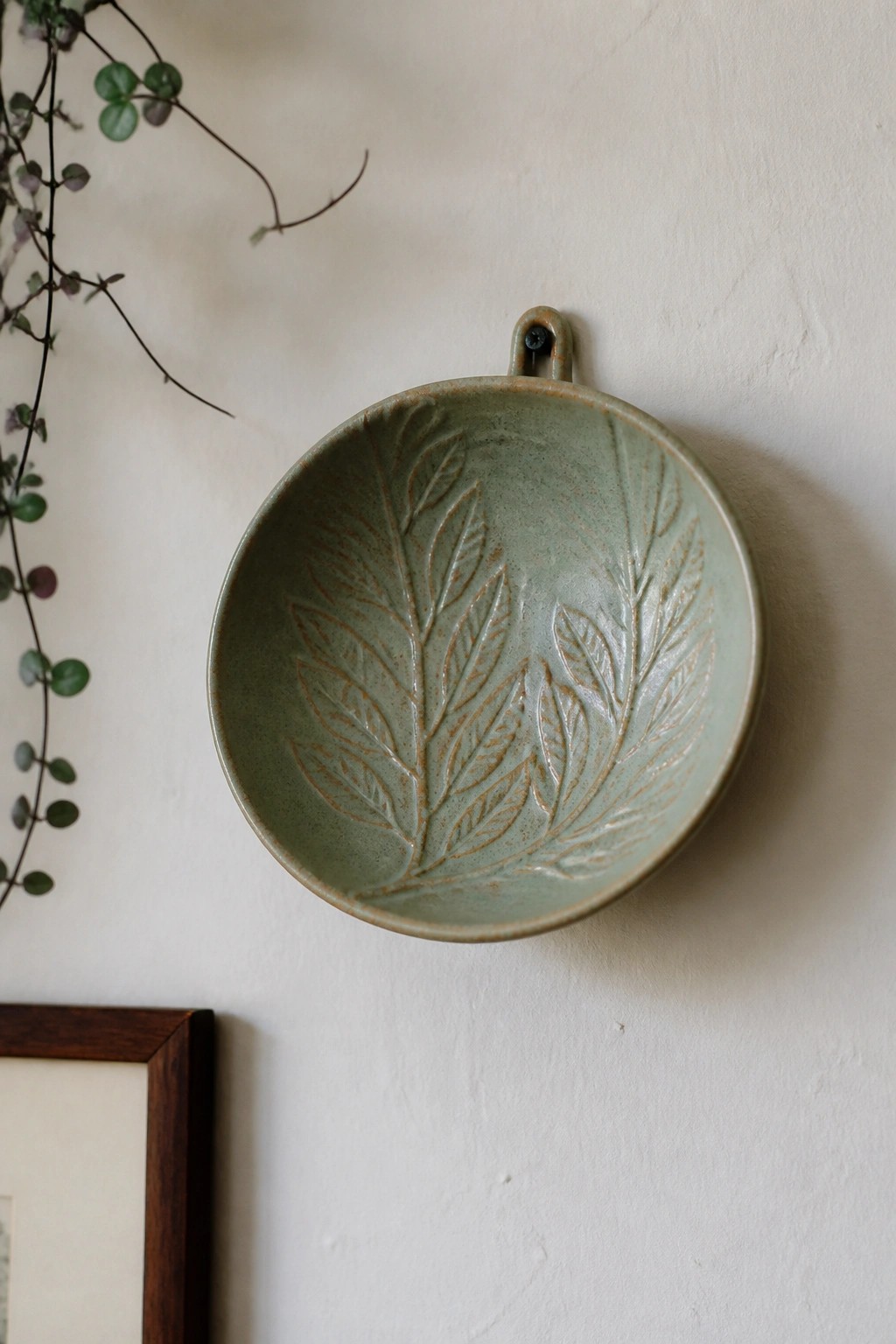 Sage green ceramic bowl with leaf patterns hanging on white wall
