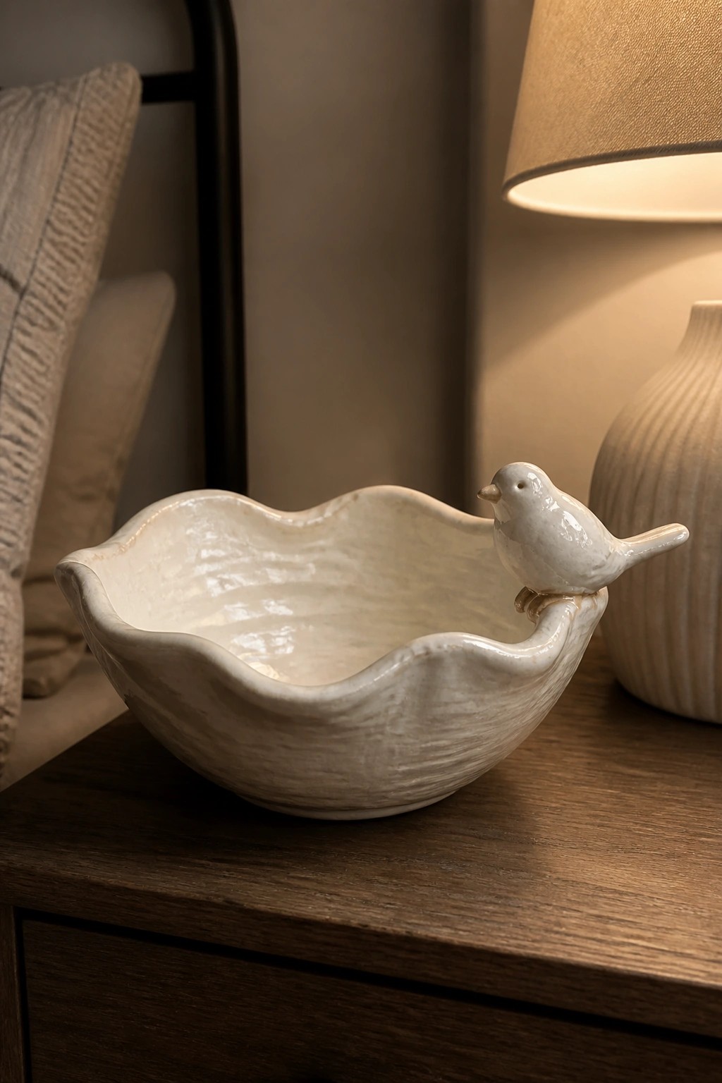 Cream ceramic bowl with bird figurine on rim atop wooden table near lamp