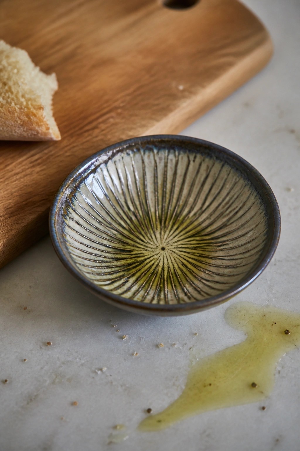 Small ceramic bowl with sunburst pattern and oil beside spill on marble near bread
