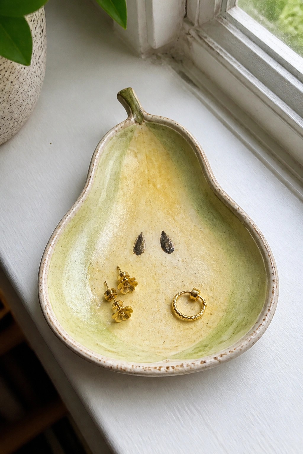 Pear-shaped ceramic dish holding gold flower earrings and hoop on windowsill