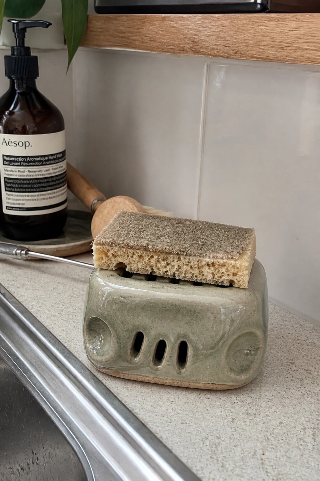 Sponge on green ceramic holder beside Aesop bottle near kitchen sink
