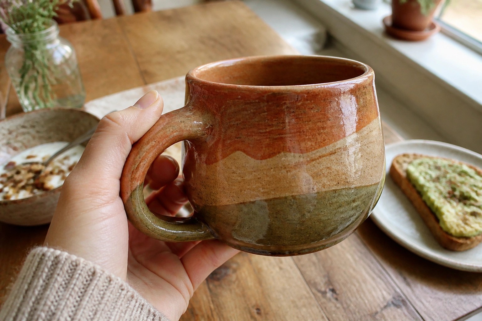 Hand in green knit sweater holding swirled ceramic mug over wooden table