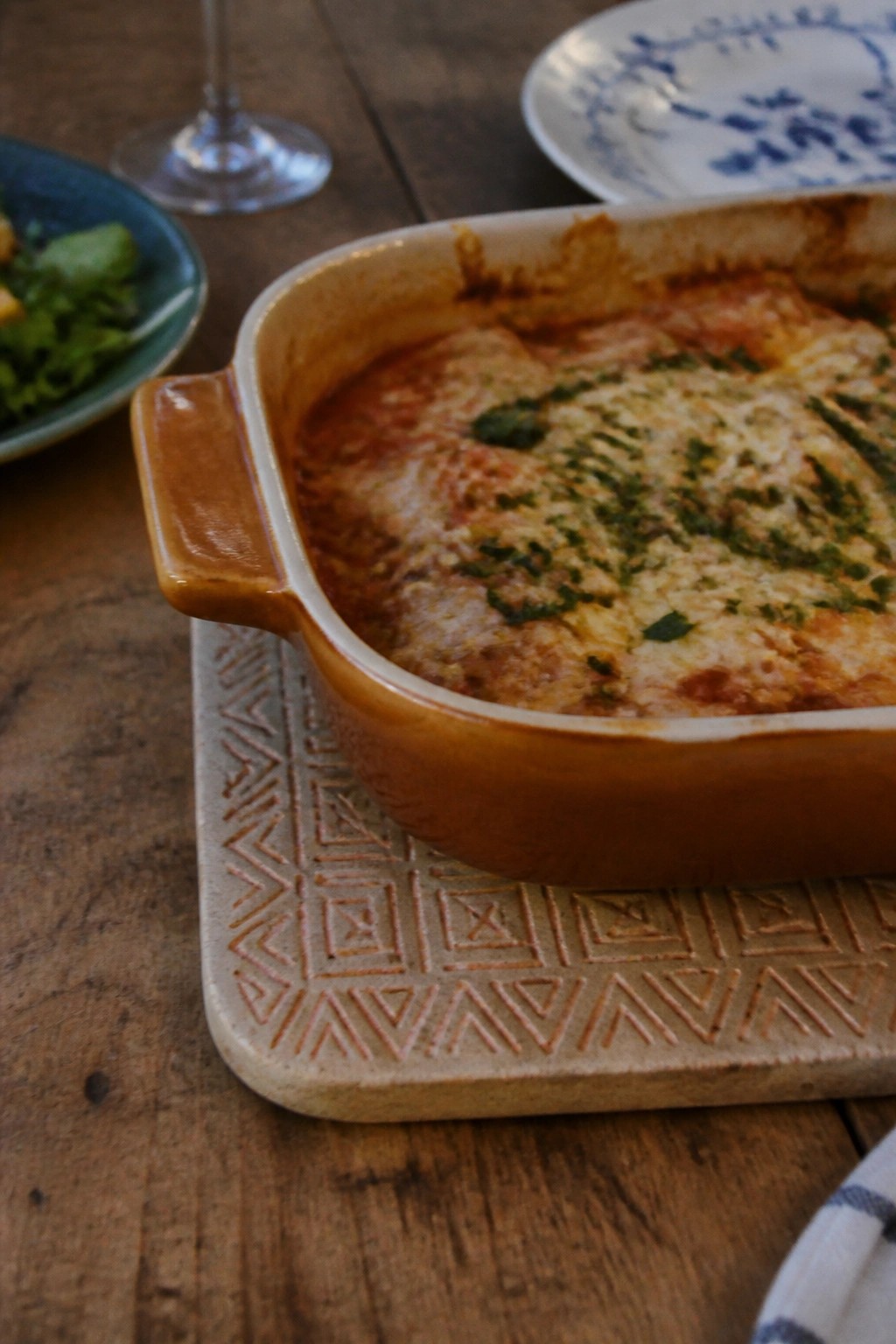 Cheesy baked pasta in orange ceramic dish on patterned trivet atop wooden table