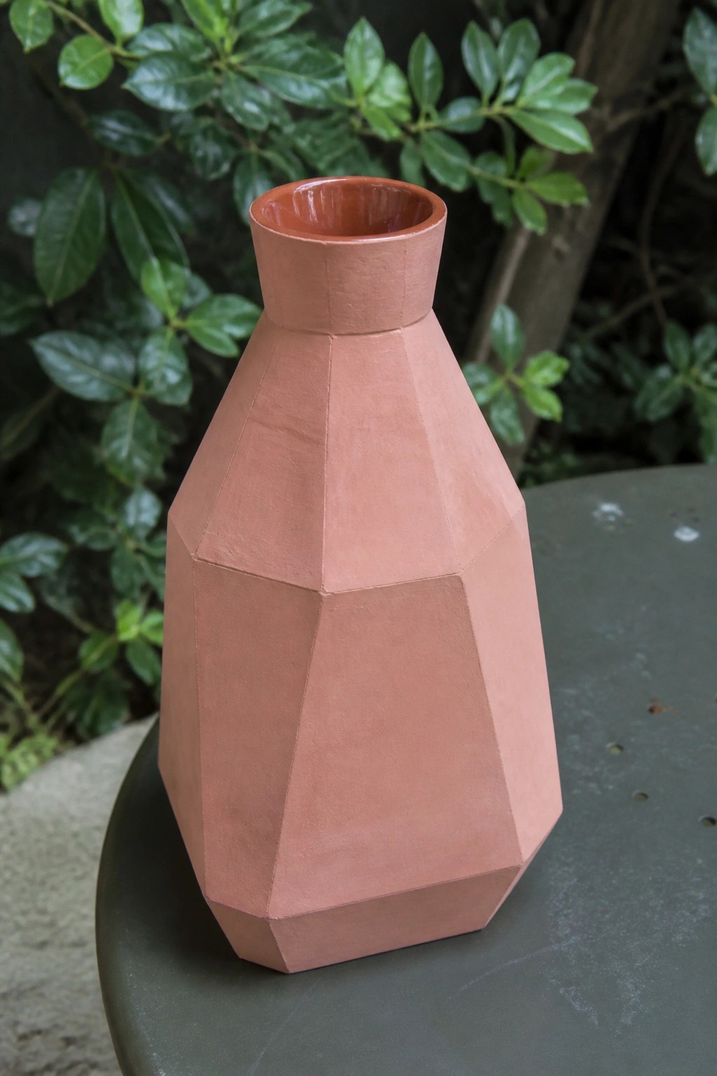 Terracotta faceted geometric vase on dark green table with leafy background.