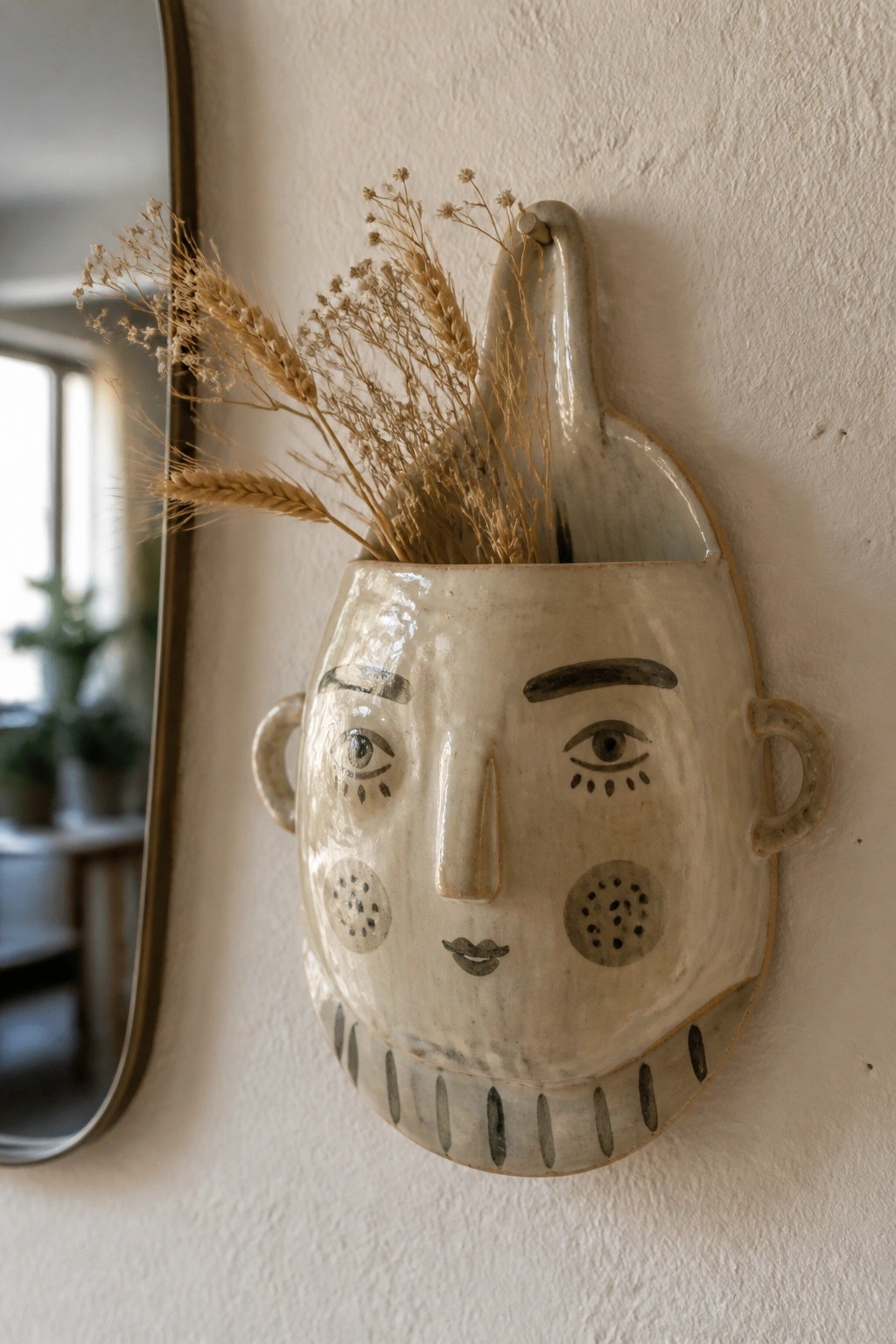 Ceramic face vase with dried grasses mounted on white wall beside mirror