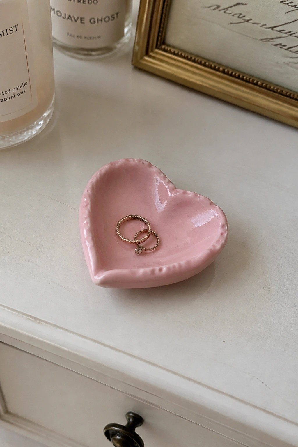 Pink heart-shaped dish holding two gold rings on white dresser with candles