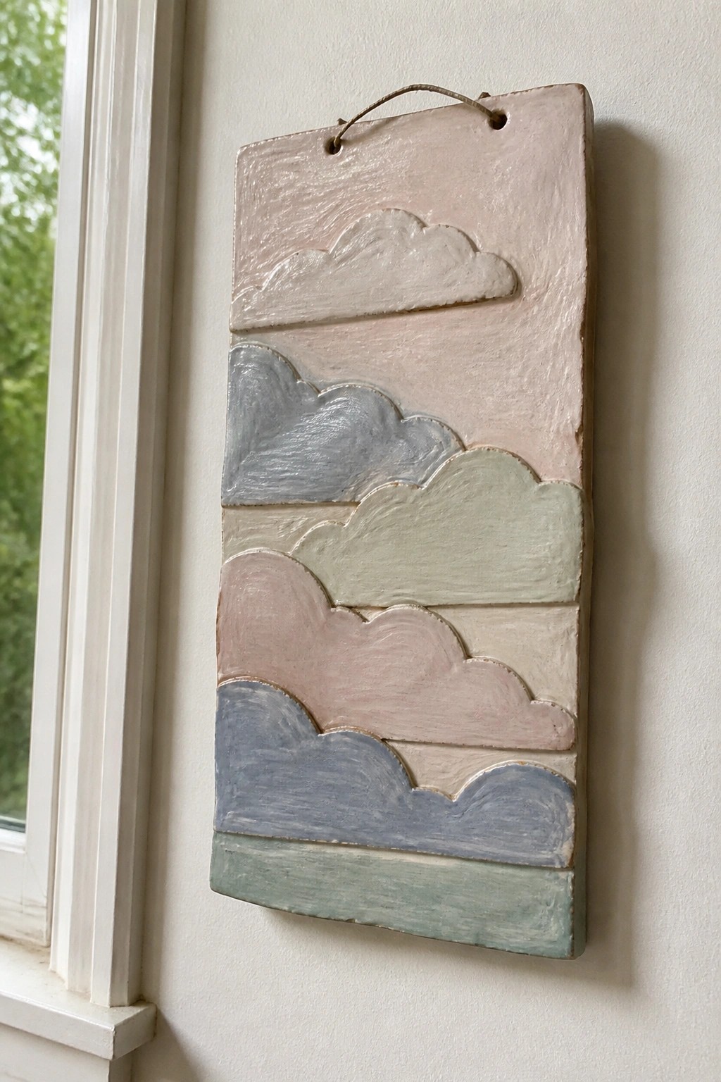 Pastel ceramic plaque with layered cloud shapes hanging beside white window frame.
