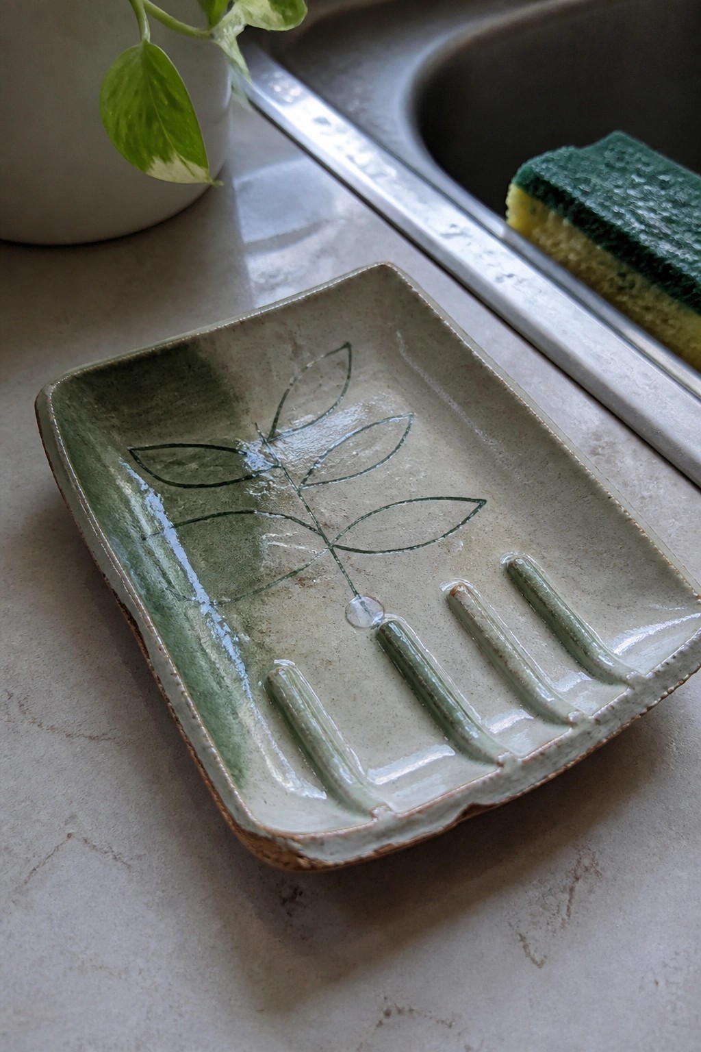 A rectangular ceramic dish with a painted green leaf design and raised ridges rests on a kitchen counter near a sink.