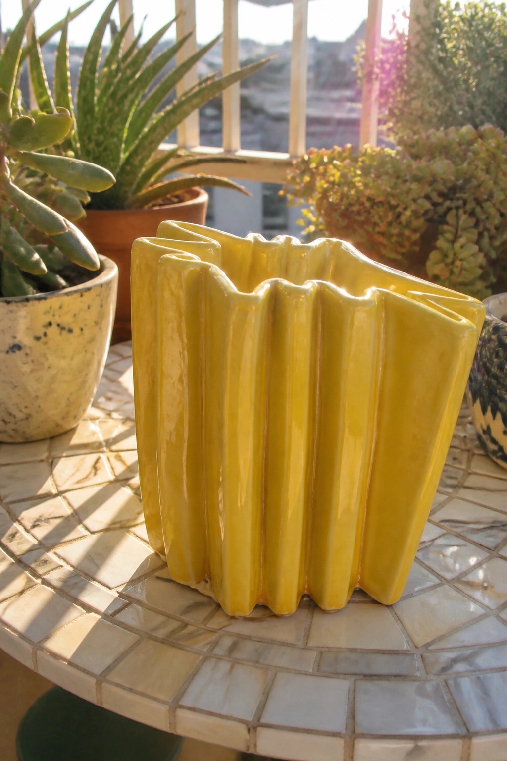 Yellow fluted ceramic vase on tiled table amid potted succulents in sunlight