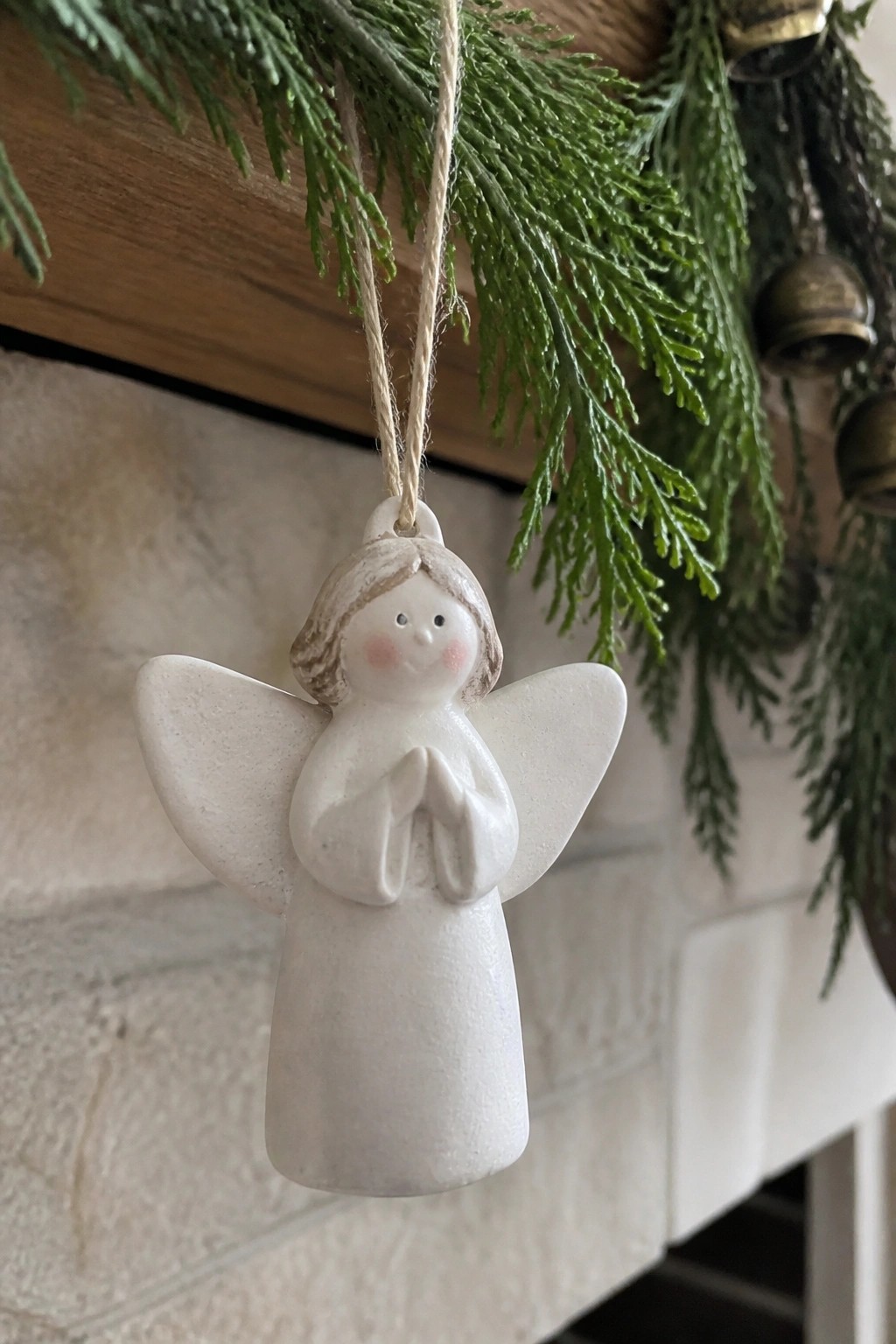 White ceramic angel ornament with wings and clasped hands hanging amid pine branches.