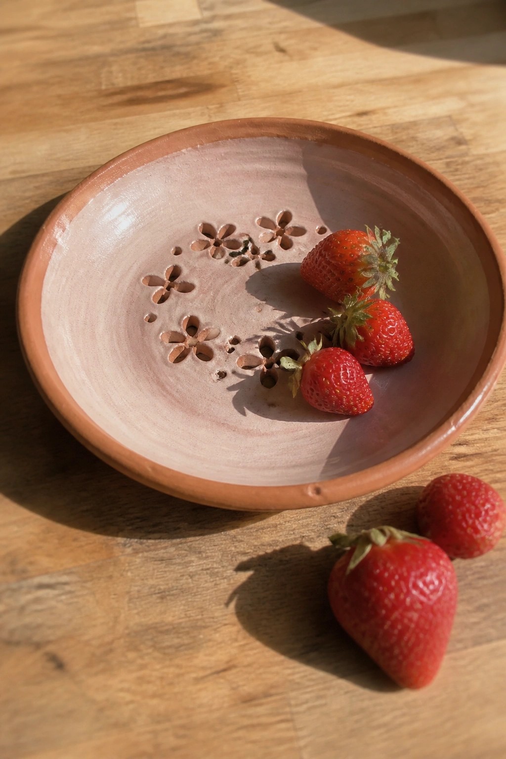 Pink pottery bowl with flower cutouts and strawberries on wooden table
