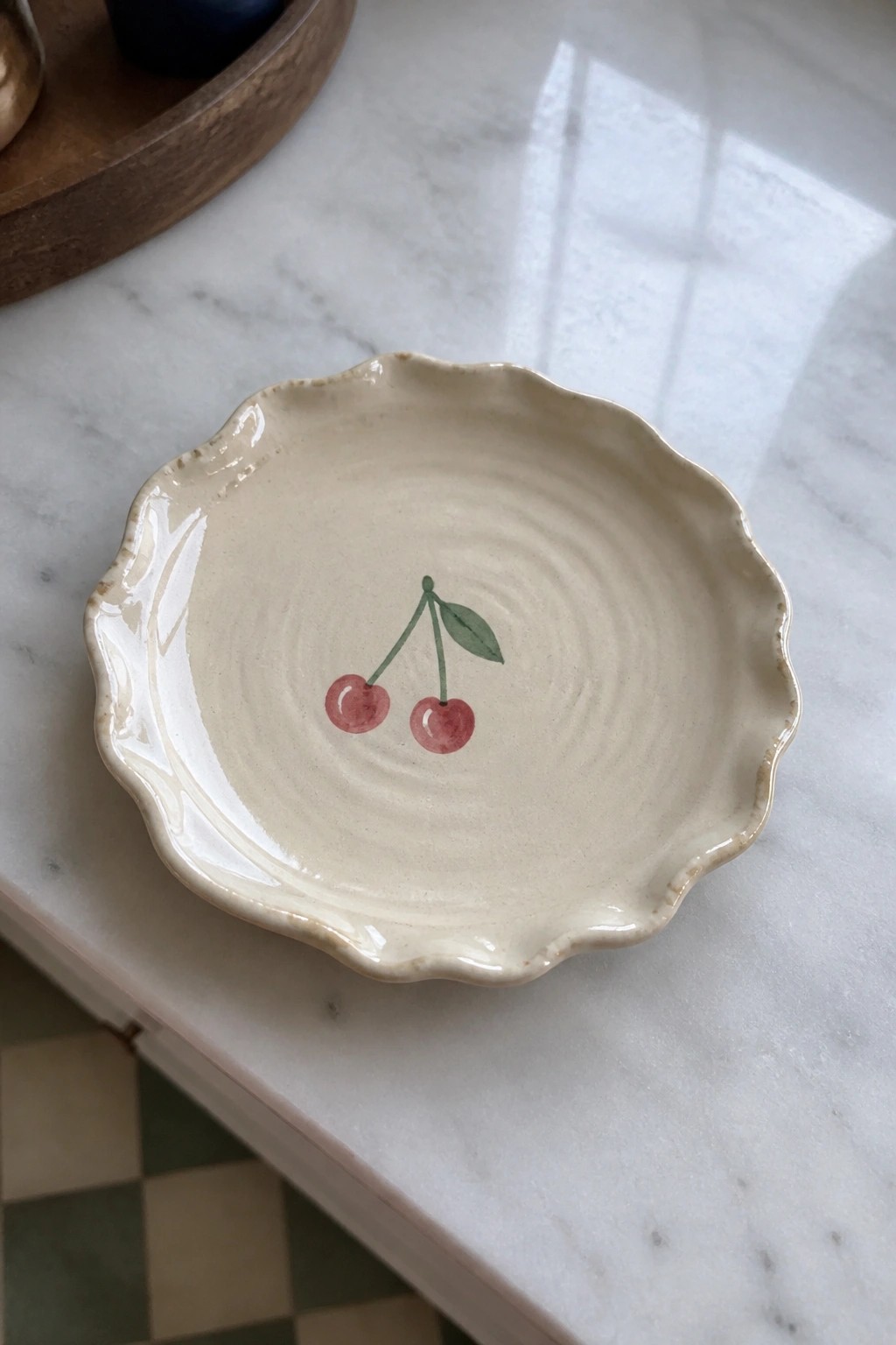 Scalloped cream ceramic dish with two painted cherries on marble surface.