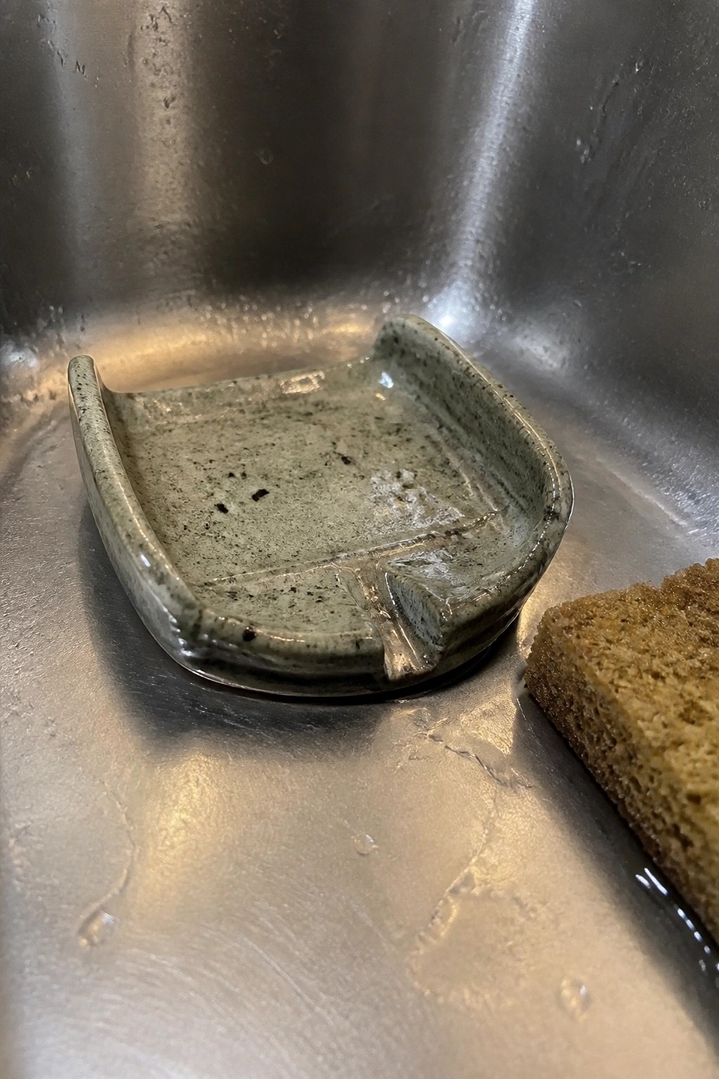 Speckled green ceramic dish in wet stainless steel sink beside bread slice