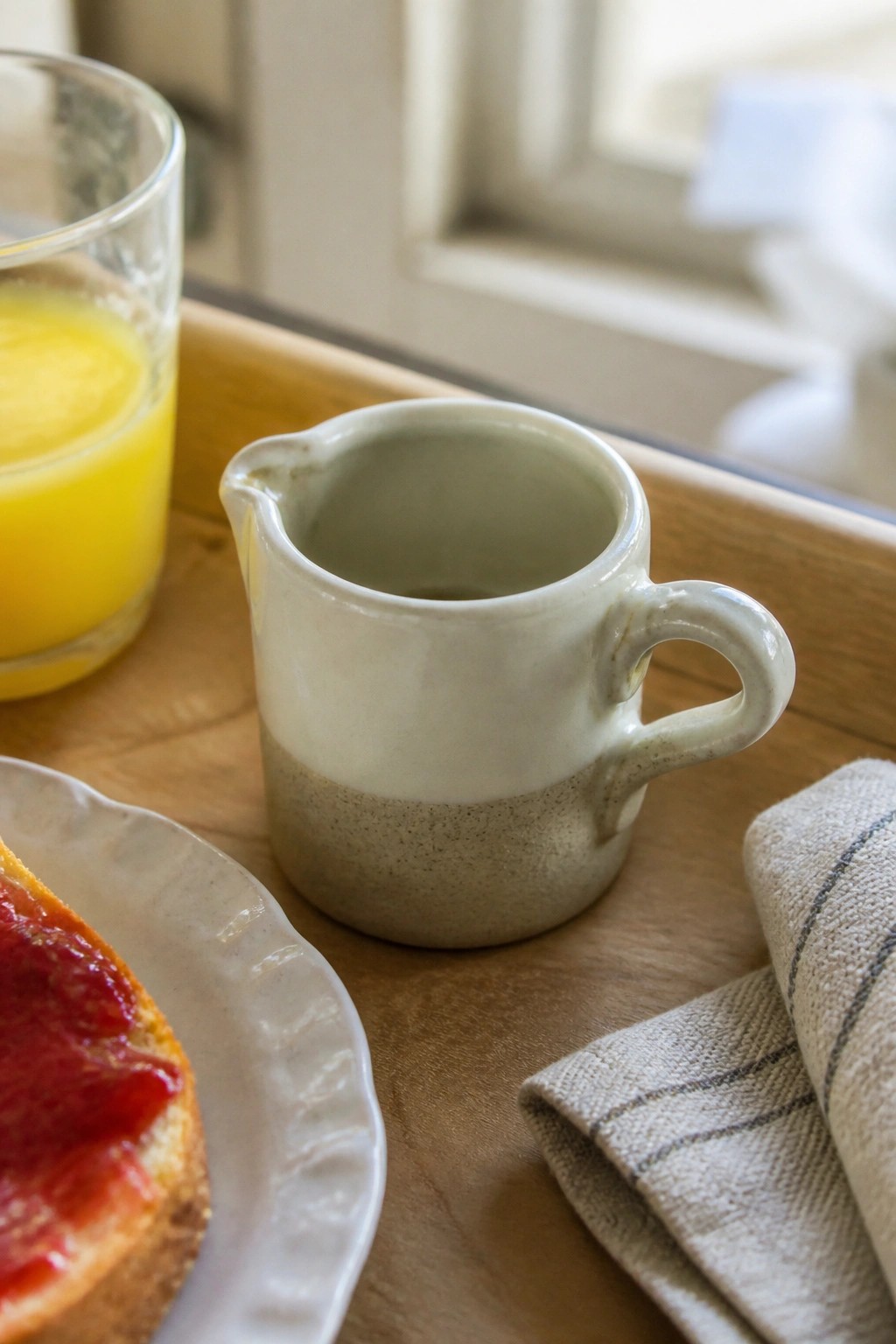 Ceramic creamer, orange juice glass, jam toast, and striped napkin on wooden tray.