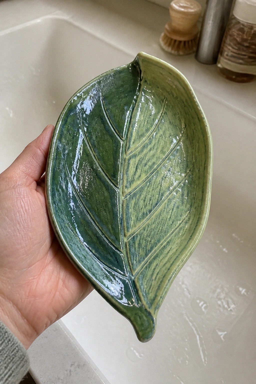 Hand holding glossy green leaf-shaped ceramic bowl over white sink.