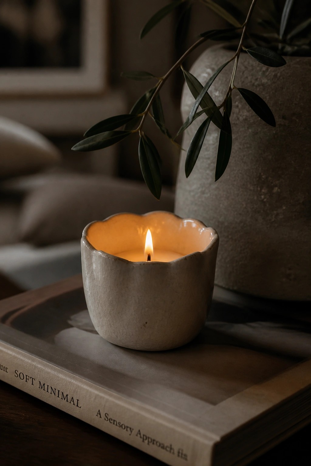 Lit candle in scalloped white ceramic holder on book beside olive branches.
