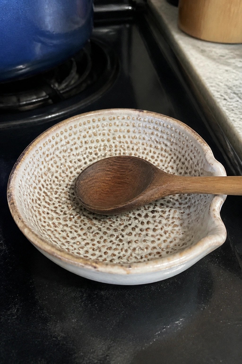Speckled ceramic bowl holding wooden spoon on black stovetop near blue pot