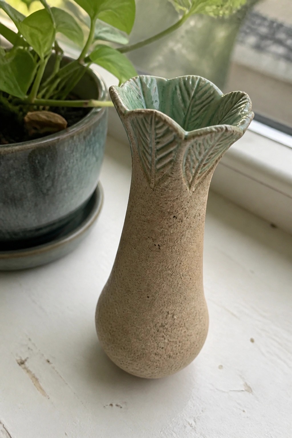 Small beige ceramic vase with green leaf-patterned rim on white windowsill beside plant