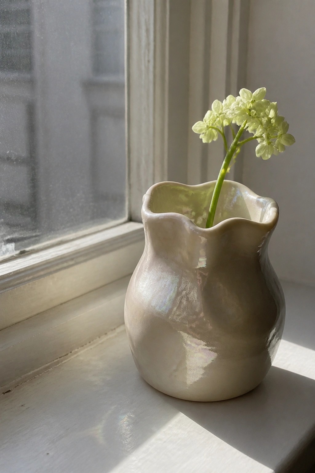 Small glossy off-white ceramic vase with wavy ruffled rim holding one light green flower stem on a windowsill.