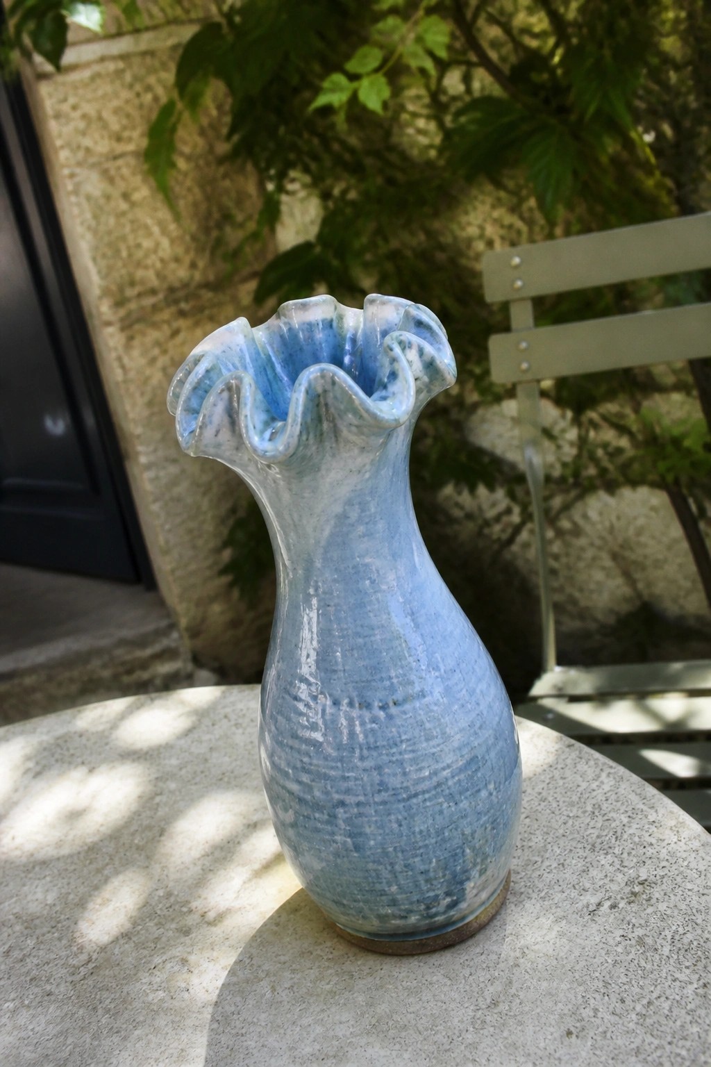 Light blue ruffled ceramic vase on sunlit stone table with green foliage behind.