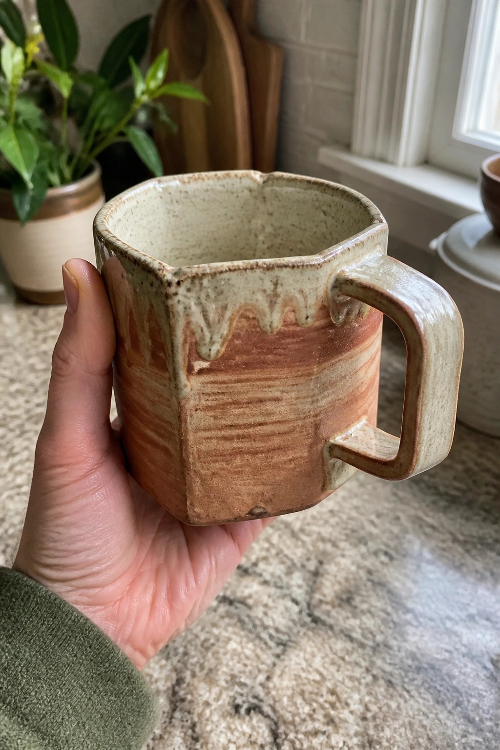 Hand in green sleeve holding faceted ceramic mug with brown drip glaze