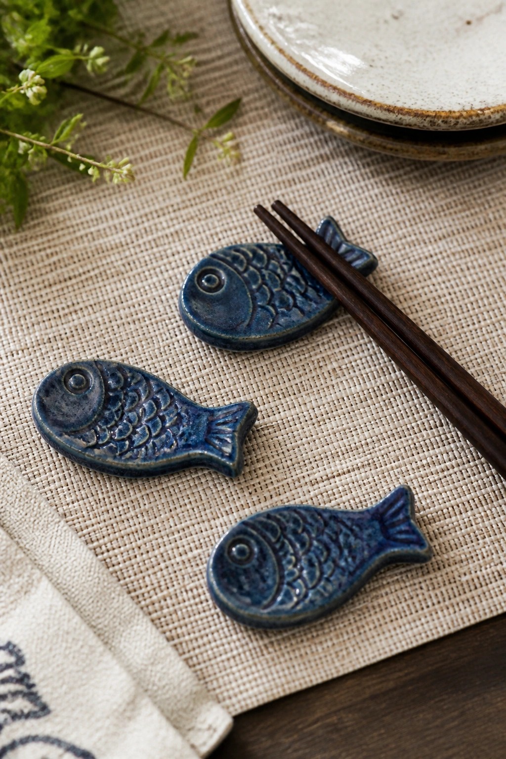 Three blue ceramic fish chopstick rests with wooden chopsticks on woven fabric beside plates