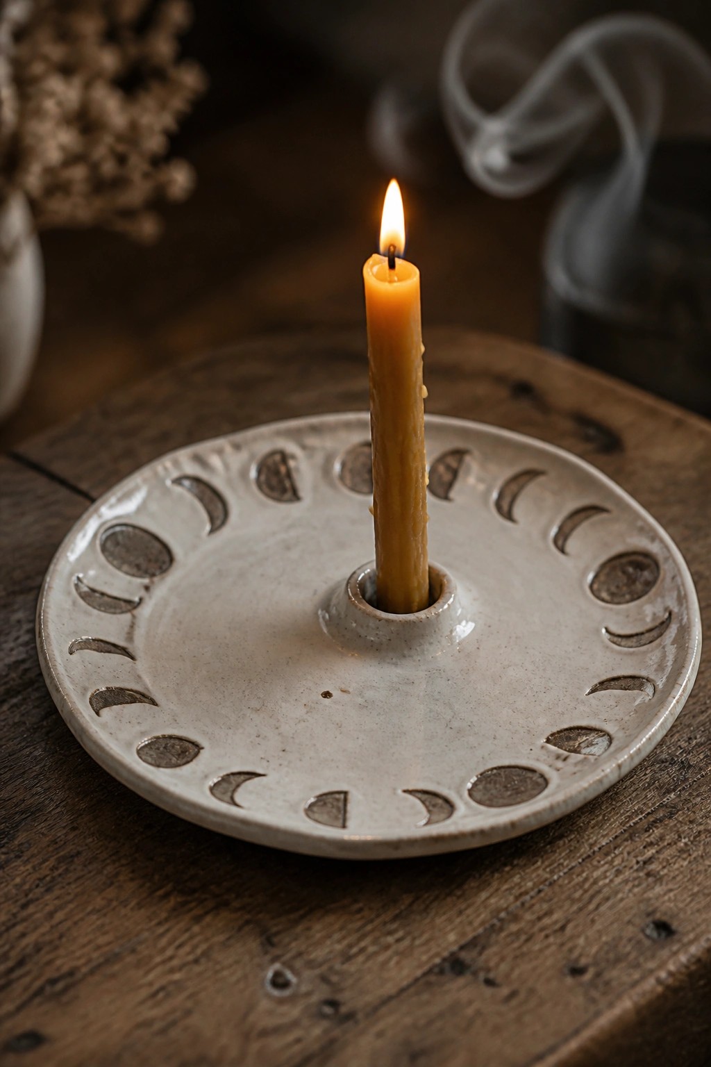 Lit yellow candle in white ceramic holder with moon phase symbols on wood.