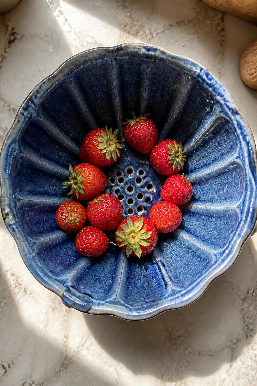 Blue ceramic colander filled with fresh red strawberries on sunlit marble.