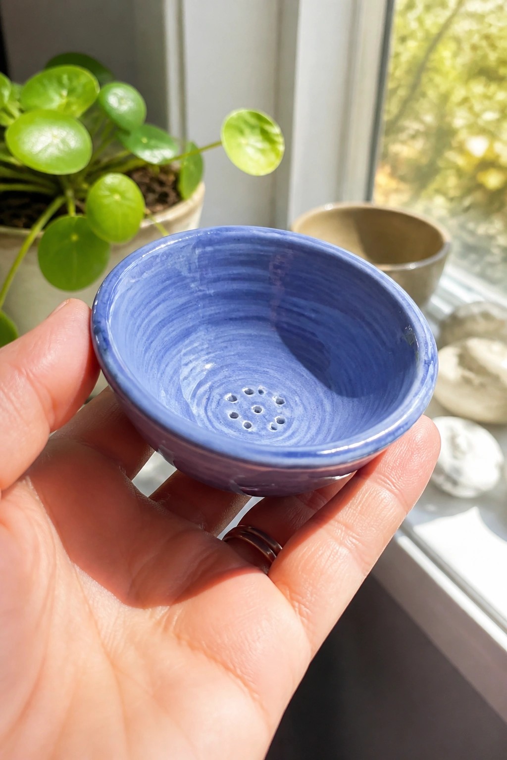 Hand holding small blue ceramic bowl with holes near window and plant.