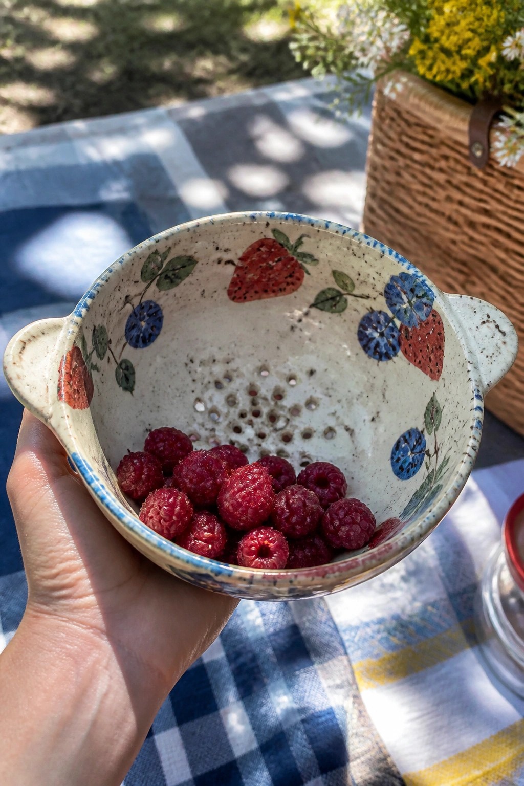 Hand holding a fruit-patterned ceramic colander filled with fresh raspberries on a picnic blanket