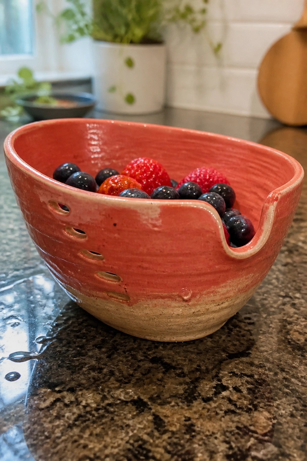 Red ceramic bowl with holes holding blueberries and raspberries on granite counter