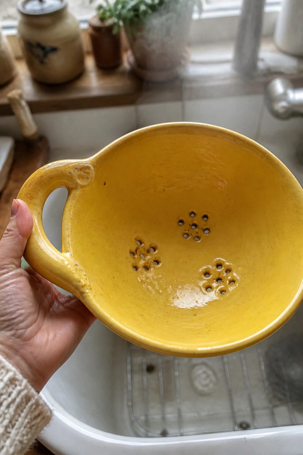 Hand in knit sweater holding yellow ceramic colander over white kitchen sink
