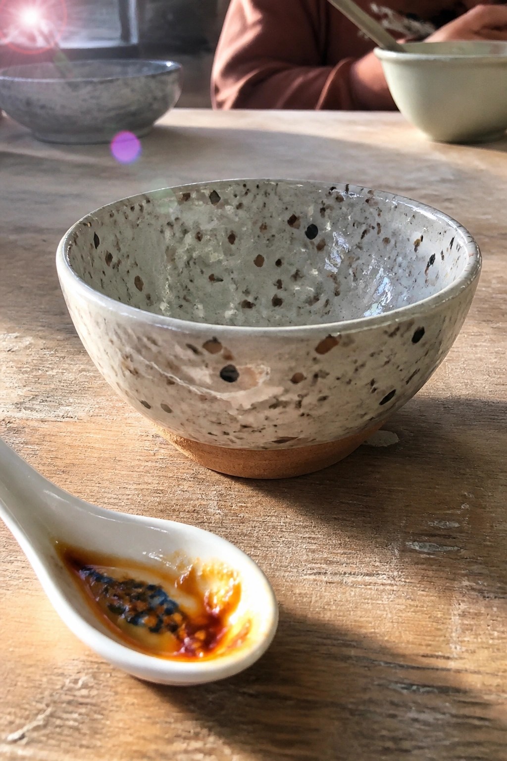 A speckled ceramic bowl on a wooden table with a white spoon holding dark sauce in front of it.
