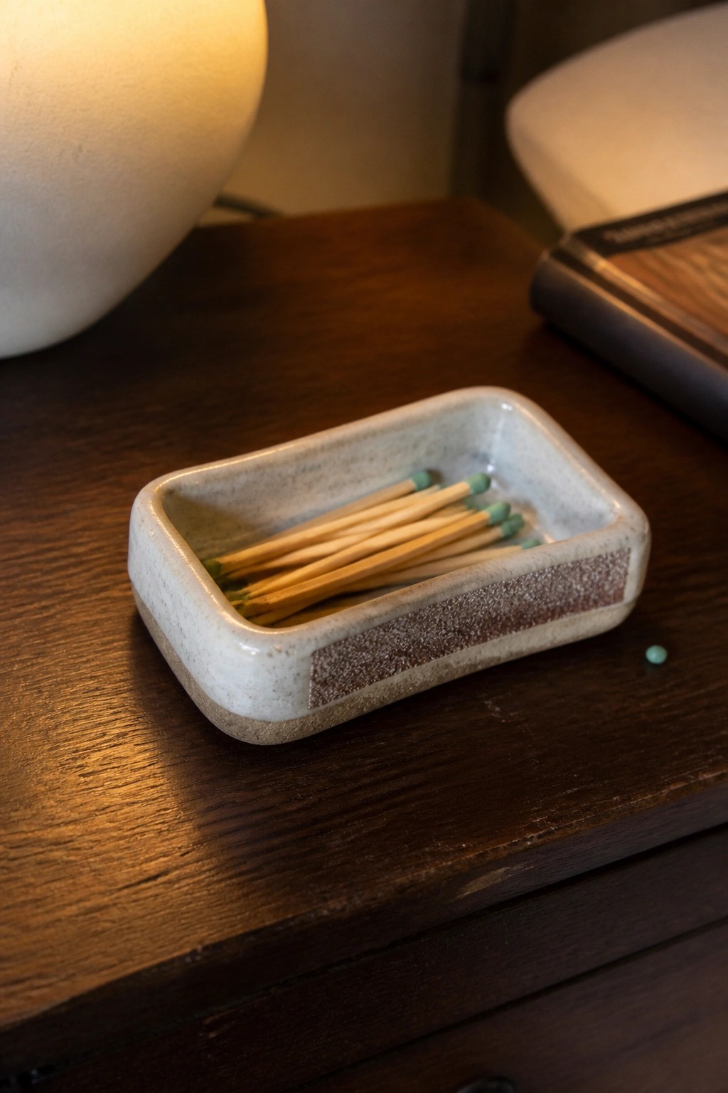 Ceramic dish holding green-tipped matches on dark wooden table near lamp.